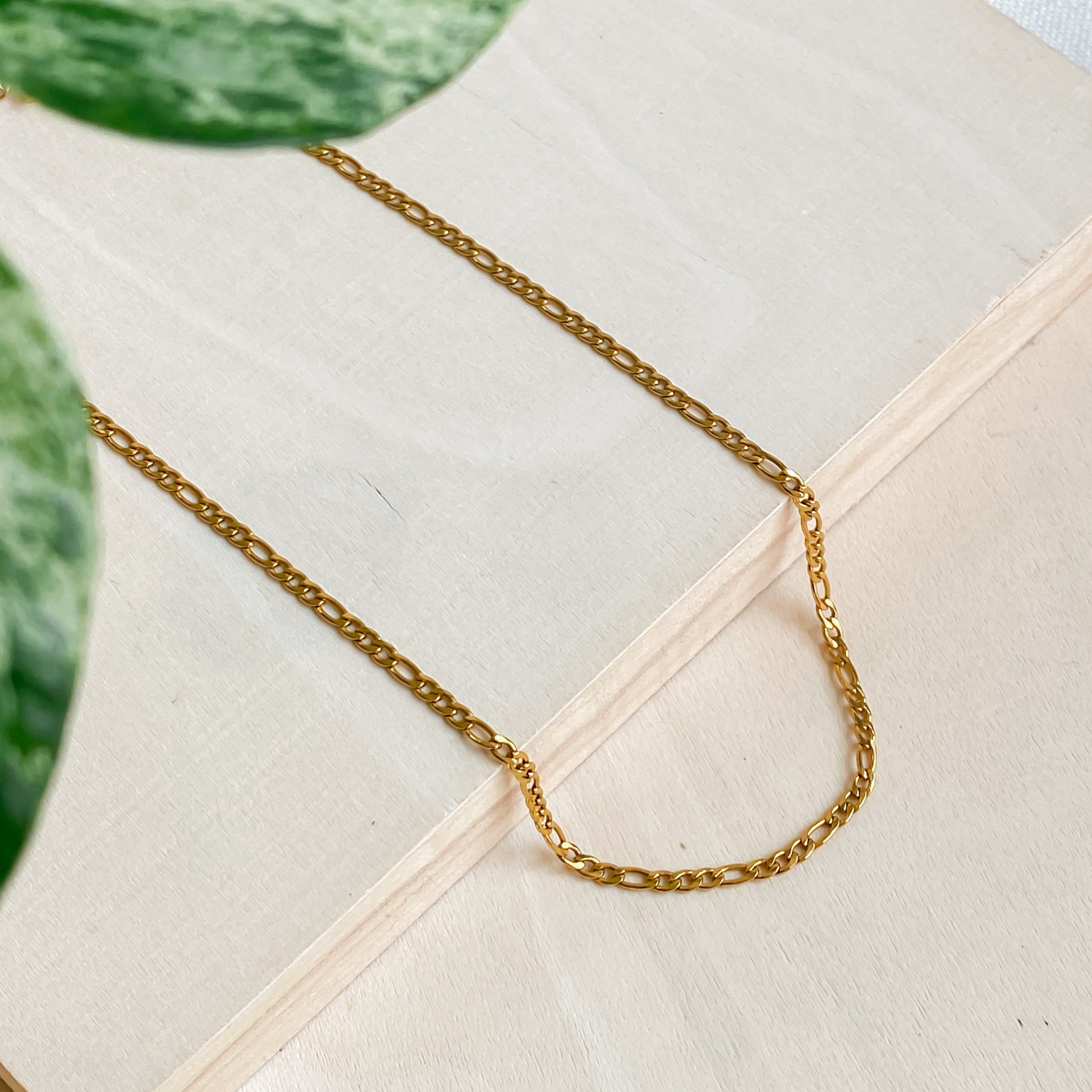 Collier Aurora en acier inoxydable doré 18K photographié à plat sur fond bois clair avec feuille verte.
