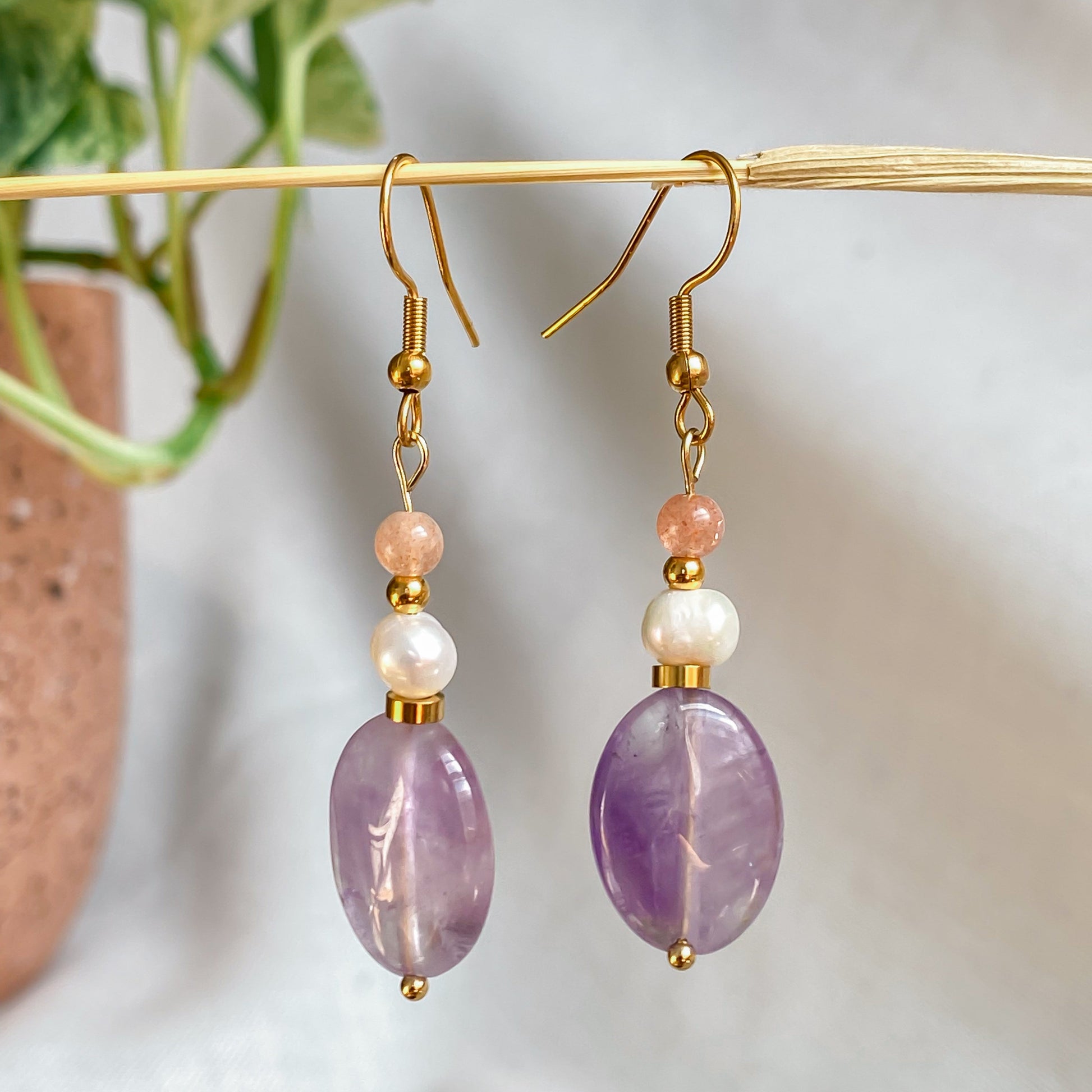 Boucles d’oreilles pendantes Lunara accrochées à une tige dorée, sur fond clair avec des feuilles vertes floutées en arrière-plan.