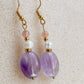 Paire de boucles d’oreilles Lunara en améthyste et perle d’eau douce, avec pierre de soleil rose, posées sur fond beige.