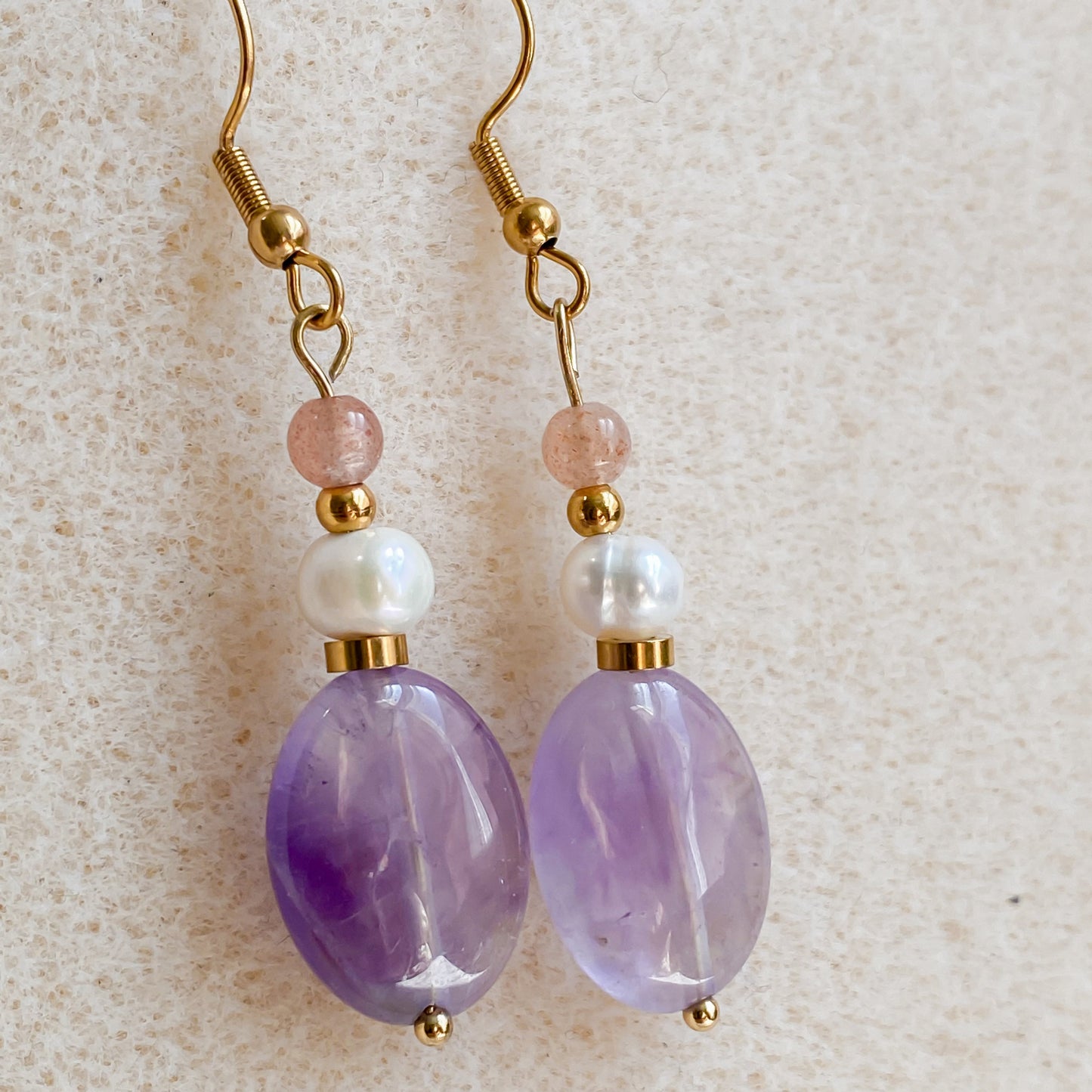 Paire de boucles d’oreilles Lunara en améthyste et perle d’eau douce, avec pierre de soleil rose, posées sur fond beige.