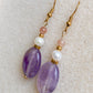 Boucles d’oreilles Lunara posées sur une surface texturée beige, mettant en valeur l’améthyste mauve et la perle d’eau douce.