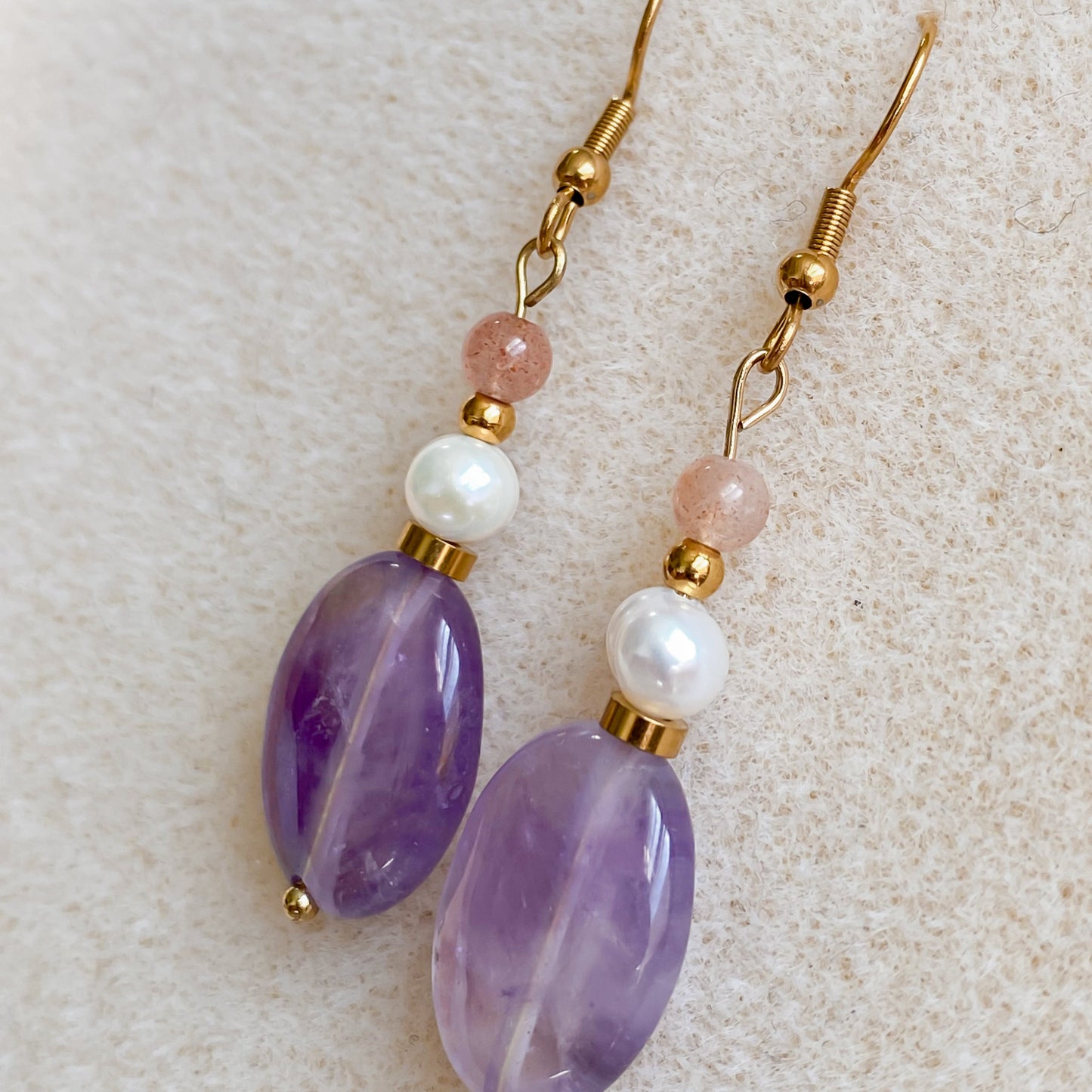 Boucles d’oreilles Lunara posées sur une surface texturée beige, mettant en valeur l’améthyste mauve et la perle d’eau douce.