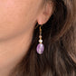 Boucles d’oreilles pendantes Lunara en améthyste, perle d’eau douce et pierre de soleil, portées à l’oreille avec crochet doré.
