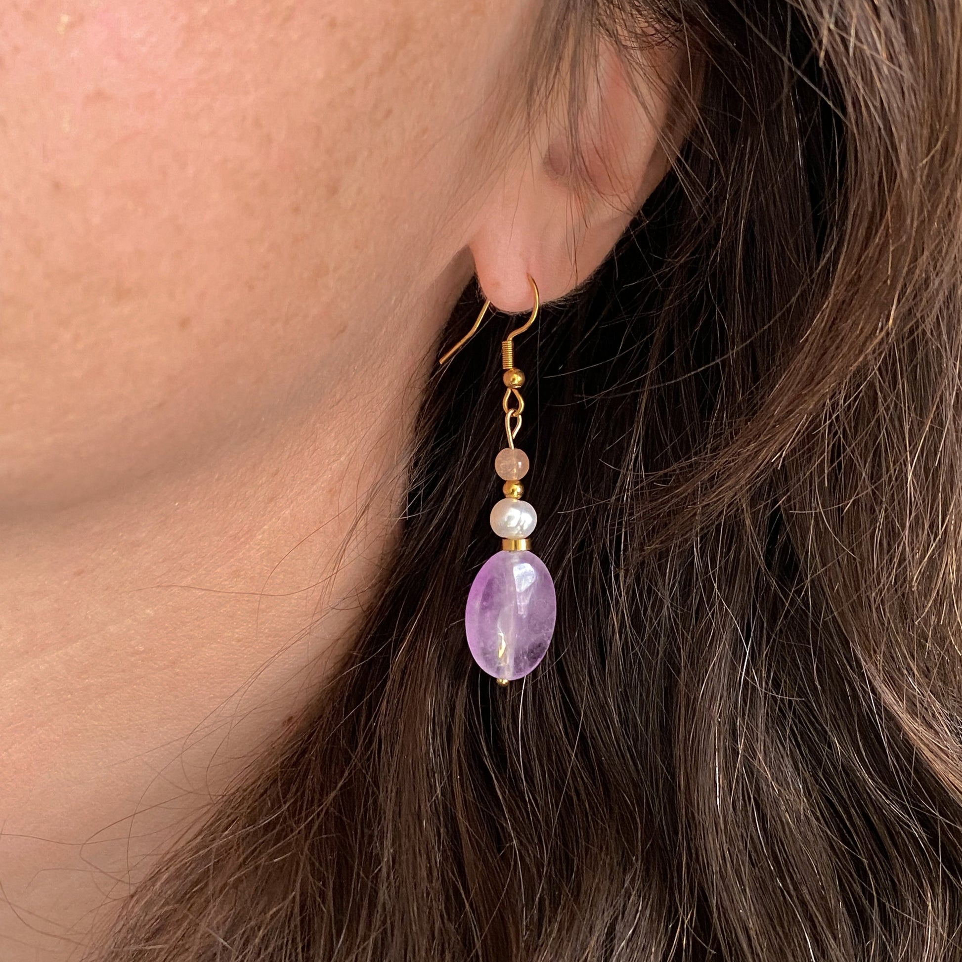 Boucles d’oreilles pendantes Lunara en améthyste, perle d’eau douce et pierre de soleil, portées à l’oreille avec crochet doré.
