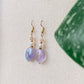 Boucles d’oreilles pendantes Lunara en améthyste avec pierres naturelles, sur fond beige, avec des feuilles floutées au premier plan.