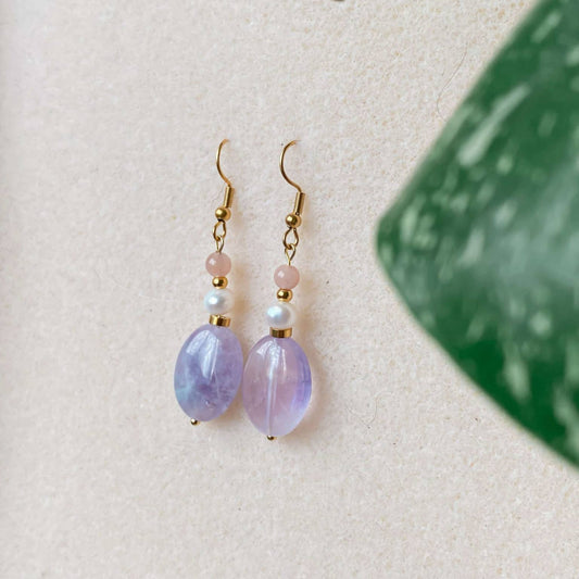 Boucles d’oreilles pendantes Lunara en améthyste avec pierres naturelles, sur fond beige, avec des feuilles floutées au premier plan.